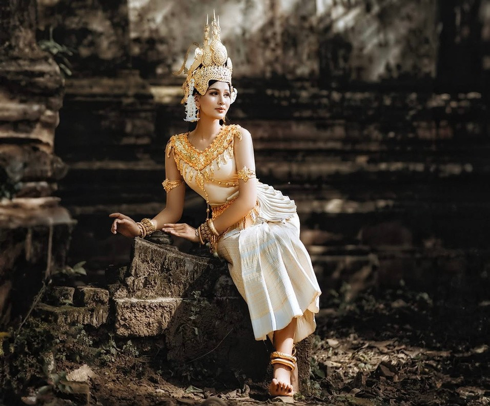 Thiếu nữ Khmer trong trang phục truyền thống với chiếc váy quấn và dải khăn vắt chéo vai trong khuôn viên chùa tháp thanh bình, dưới bóng dáng của những bức tượng Phật uy nghi. (Ảnh: Chú Râu)